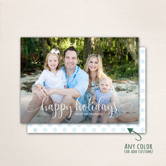Beach-inspired family holiday card with “Happy Holidays” in graceful white script. The design highlights a family of four seated by the shore with lush green trees in the background and an optional snowflake-patterned back.