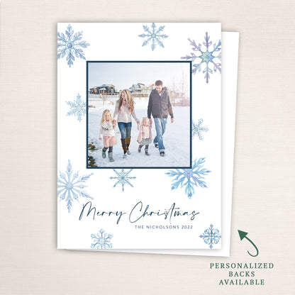 “Merry Christmas” snowflake photo card from Halo Design Shop featuring a centered family photo framed in navy blue, surrounded by watercolor snowflakes in cool winter tones. Includes customizable text, color options, and optional personalized backs.