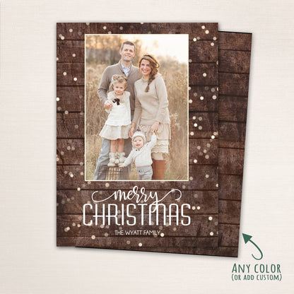 Vertical “Merry Christmas” photo card featuring a family of four on a rustic wood background with soft snowflake accents. The design includes modern white lettering and a customizable color option.