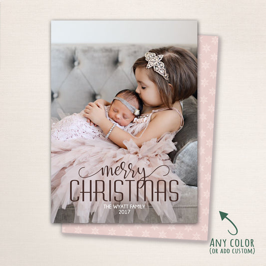 Vertical Christmas photo card with a soft blush background and elegant “Merry Christmas” script overlay. Features a little girl lovingly holding her newborn sister on a tufted couch, with an optional snowflake-patterned back.