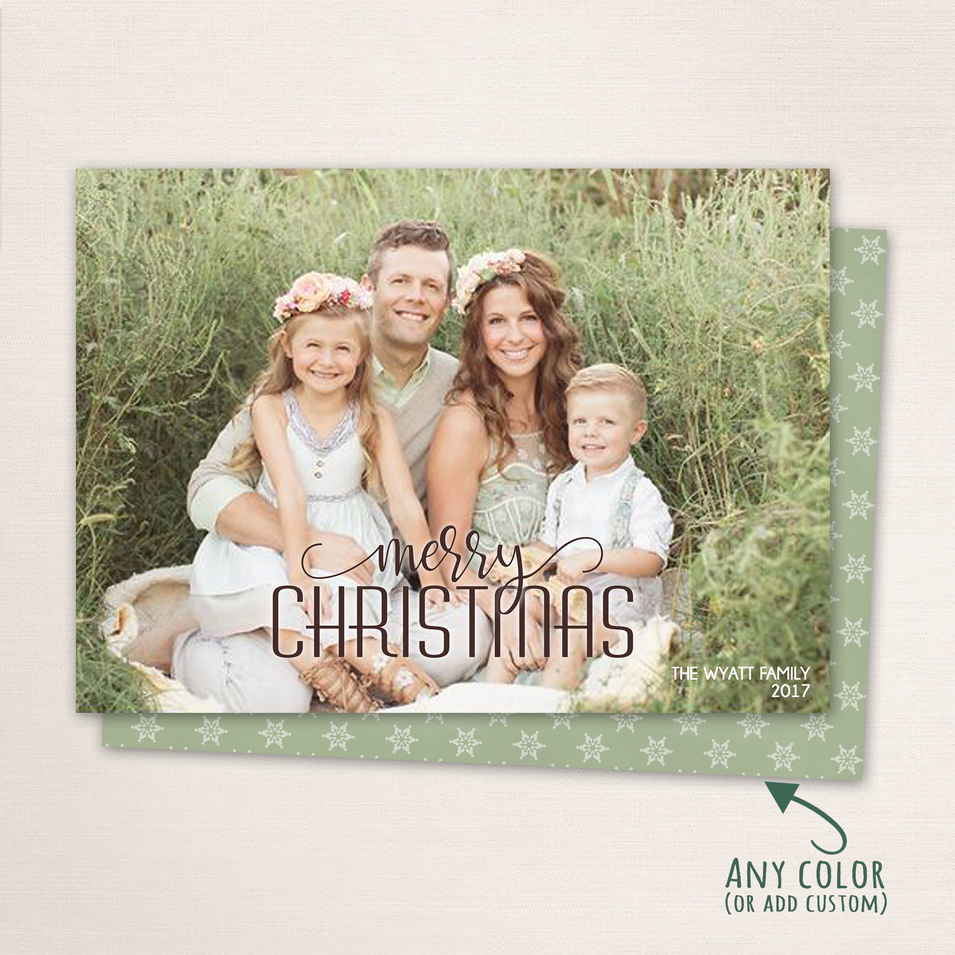 Horizontal holiday photo card featuring a family of four sitting outdoors among green foliage. The design includes “Merry Christmas” in elegant script with customizable colors and a matching snowflake-patterned back.
