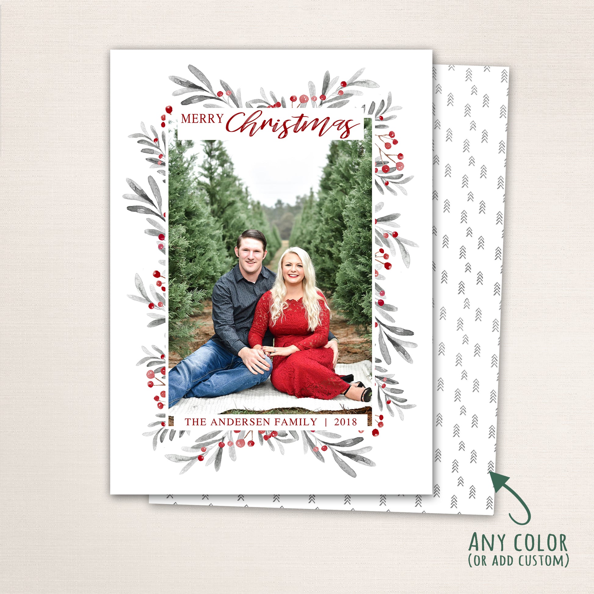 Vertical Merry Christmas photo card featuring a couple sitting among evergreen trees, surrounded by a watercolor berry and leaf wreath border with customizable family name and year and a tiny tree pattern back.