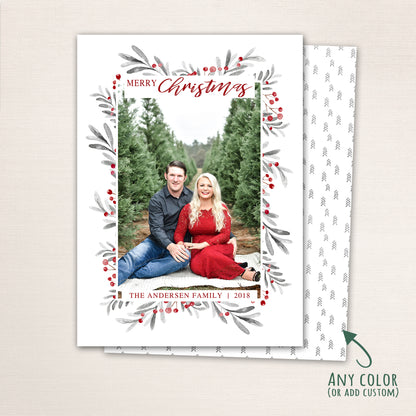 Vertical Merry Christmas photo card featuring a couple sitting among evergreen trees, surrounded by a watercolor berry and leaf wreath border with customizable family name and year and a tiny tree pattern back.