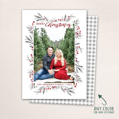 Vertical Merry Christmas photo card featuring a couple sitting among evergreen trees, surrounded by a watercolor berry and leaf wreath border with customizable family name and year and a plaid back.