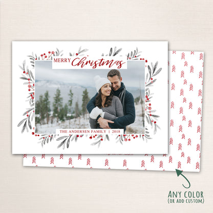 Horizontal Merry Christmas photo card featuring a couple embracing in the snow, surrounded by a watercolor berry and leaf wreath border with customizable family name and year and a mini tree pattern on the bac.