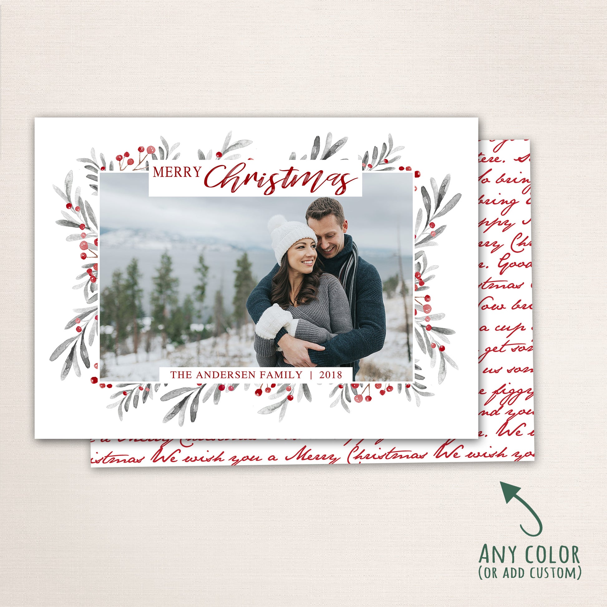 Horizontal Merry Christmas photo card featuring a couple embracing in the snow, surrounded by a watercolor berry and leaf wreath border with customizable family name and year and a Christmas carol written in script on the back.