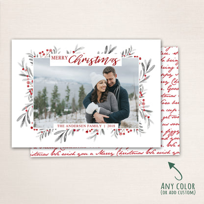 Horizontal Merry Christmas photo card featuring a couple embracing in the snow, surrounded by a watercolor berry and leaf wreath border with customizable family name and year and a Christmas carol written in script on the back.