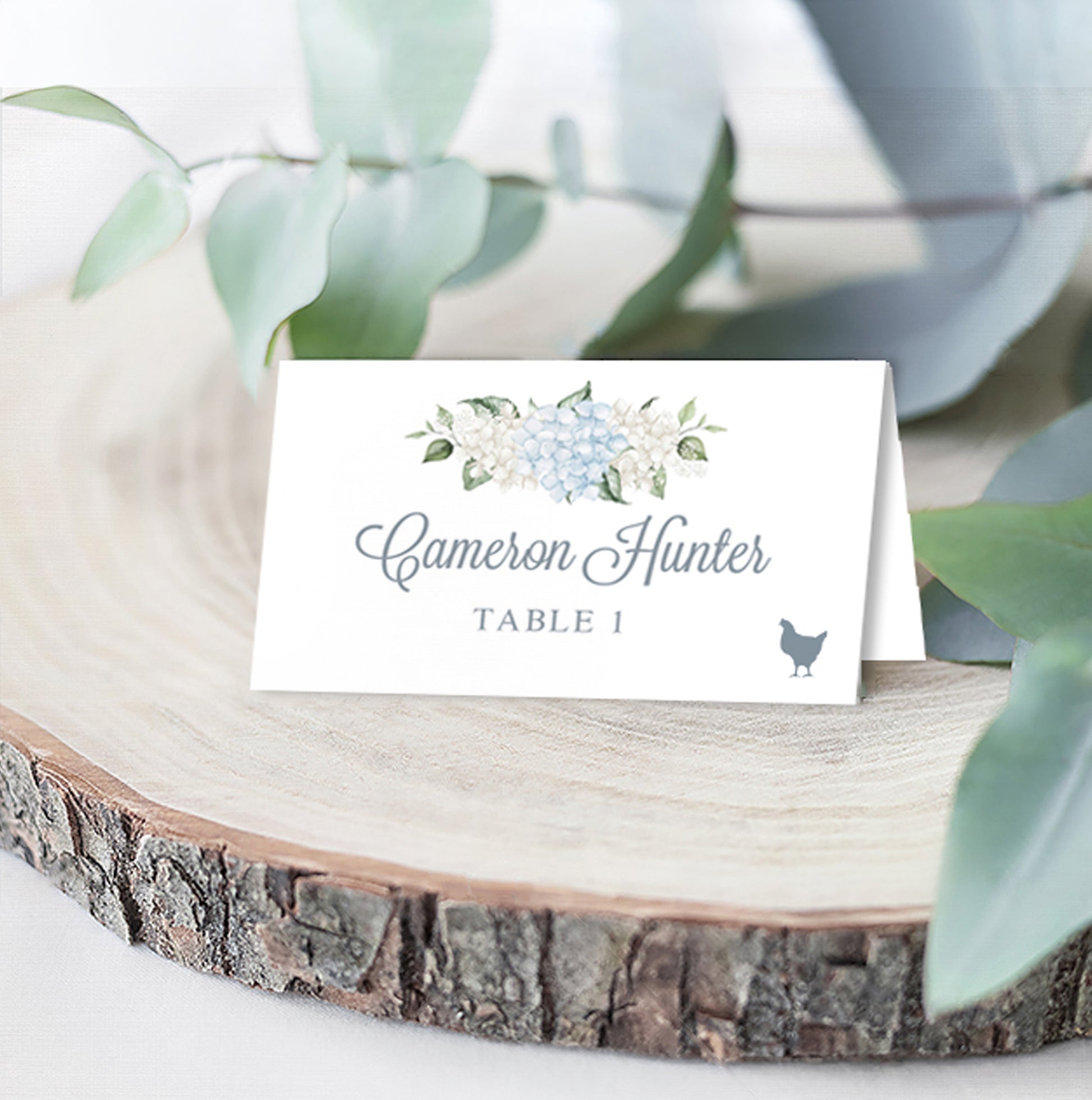 Folded wedding escort card with blue and white hydrangeas above elegant calligraphy that the guest name, table assignment, and chicken icon for their selected meal choice, styled on a wooden slice with eucalyptus leaves.