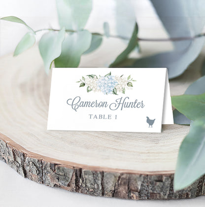 Folded wedding escort card with blue and white hydrangeas above elegant calligraphy that the guest name, table assignment, and chicken icon for their selected meal choice, styled on a wooden slice with eucalyptus leaves.