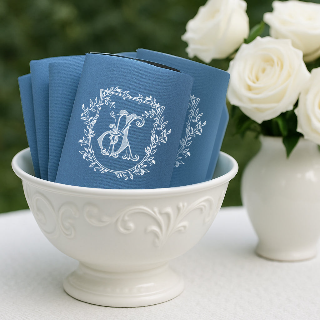 Stack of personalized navy blue wedding can cooler koozies featuring a white floral wreath monogram, displayed in a white ceramic bowl with white roses in the background.