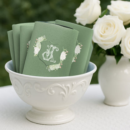 Stack of sage green personalized wedding can cooler koozies with a floral crest monogram, styled in a white bowl beside white roses for an elegant reception display.