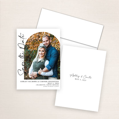 Modern photo save the date card featuring a vertical layout with an arched photo of a couple surrounded by autumn leaves. Elegant black script reads “Save the Date” along the side, with wedding details printed below. The matching back design includes their names and wedding date in minimalist black script.