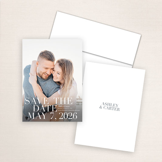 Modern photo save the date card featuring a couple embracing at the beach. The design showcases bold serif text reading “Save the Date – May 7, 2026,” with minimalist wedding details and a matching navy envelope.