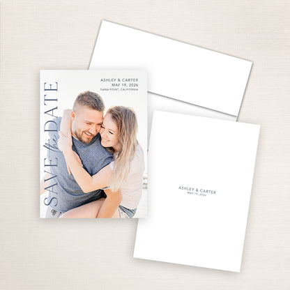 Vertical photo save the date card featuring a smiling couple embracing on the beach with elegant navy text reading “SAVE the DATE,” and wedding details for Ashley and Carter in Dana Point, California.