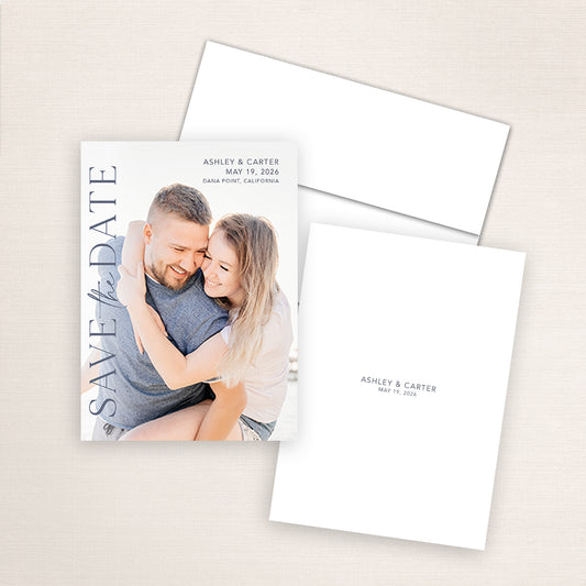 Vertical photo save the date card featuring a smiling couple embracing on the beach with elegant navy text reading “SAVE the DATE,” and wedding details for Ashley and Carter in Dana Point, California.